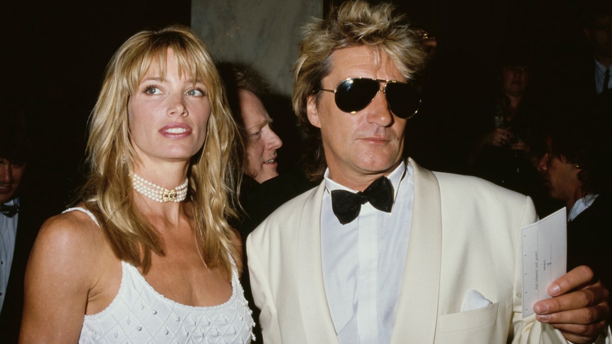 Rod Stewart in a white tuxedo poses in dark sunglasses with Kelly Emberg