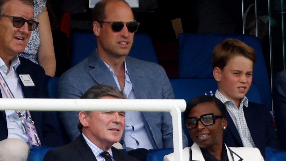 Prince William wearing sunglasses sitting in the stands next to Prince George