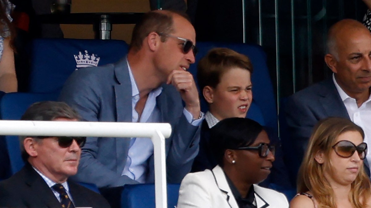 Prince William wears sunglasses and leans in to talk to Prince George in the stands