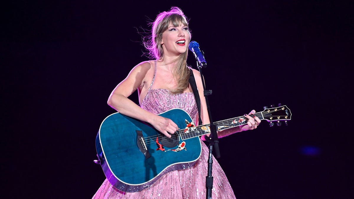 Taylor Swift in a purple dress strums a blue guitar on stage