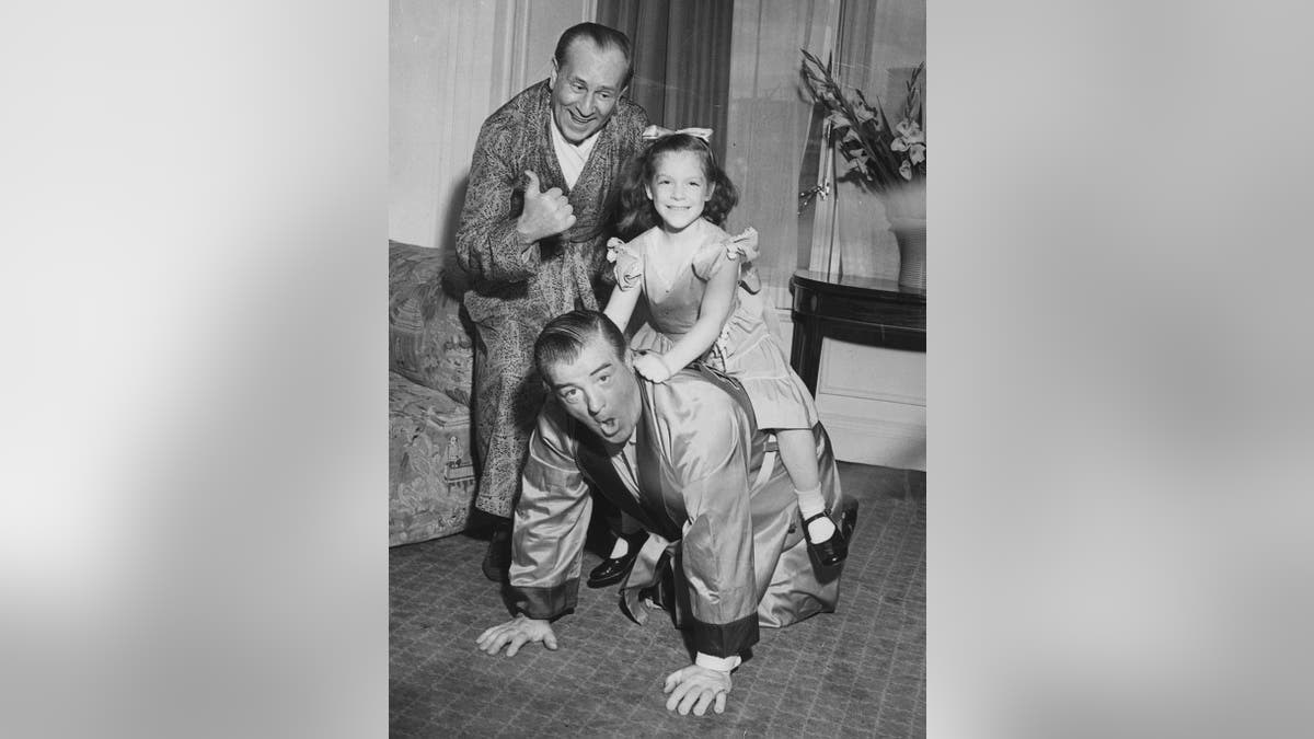 Abbott and Costello in suits and smiling at the camera as they play horse ride with daughter Chris Costello