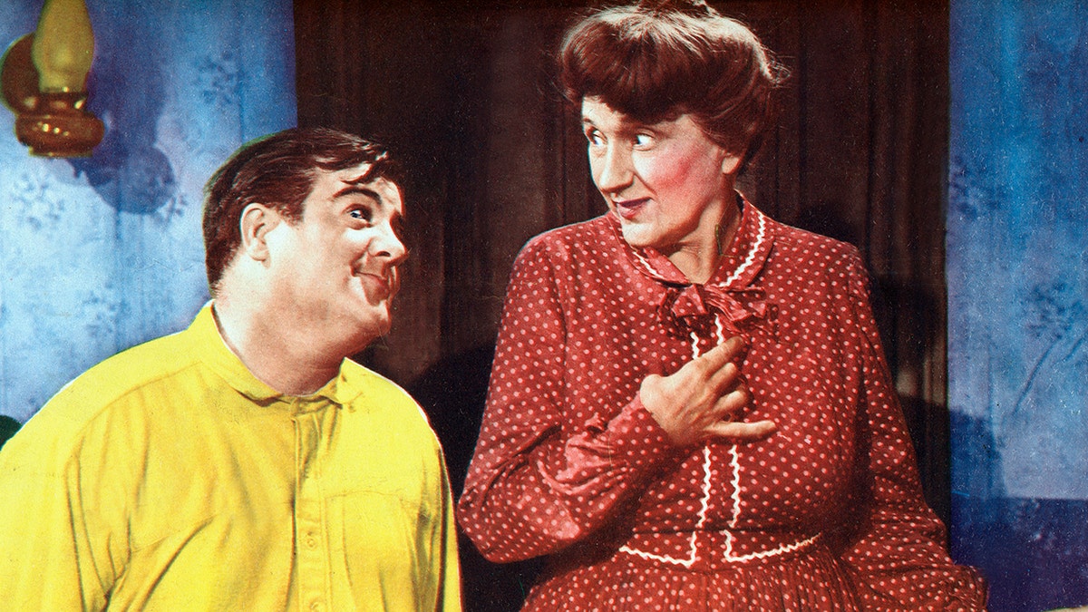 Bud Abbott wearing a polka dot burgundy dress and wig as a woman with Lou Costello wearing a yellow shirt while admiring him