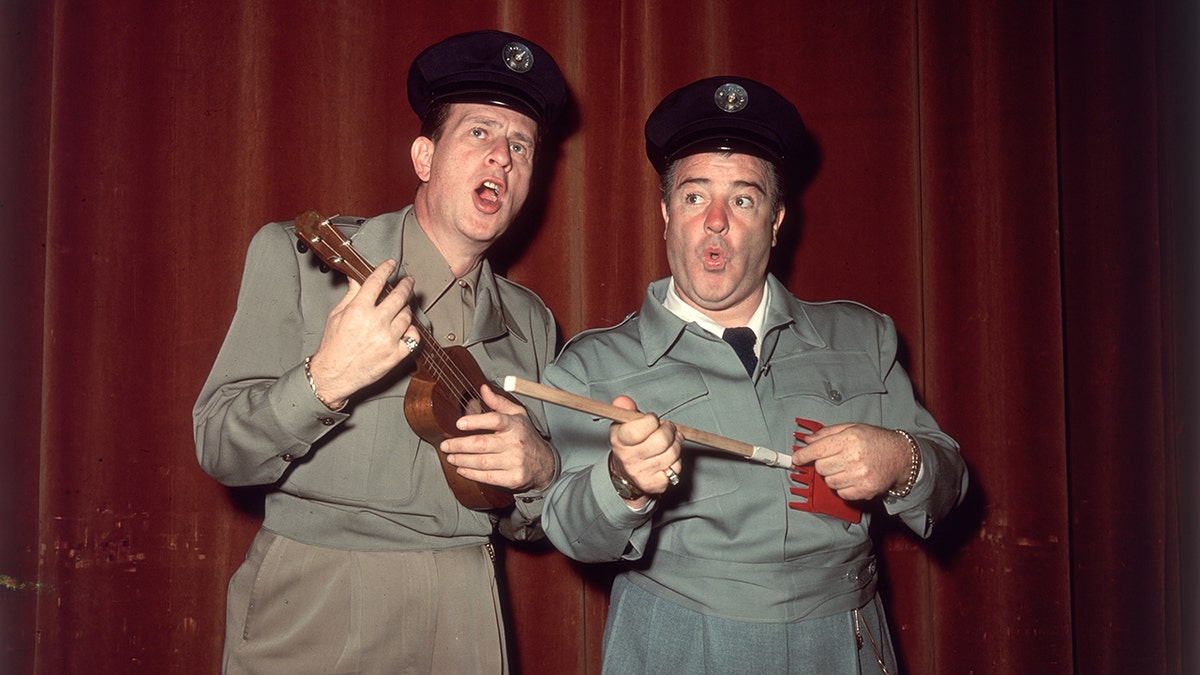 Bud Abbott holding a ukelele and singing with Lou Costello holding a gardening tool as a guitar