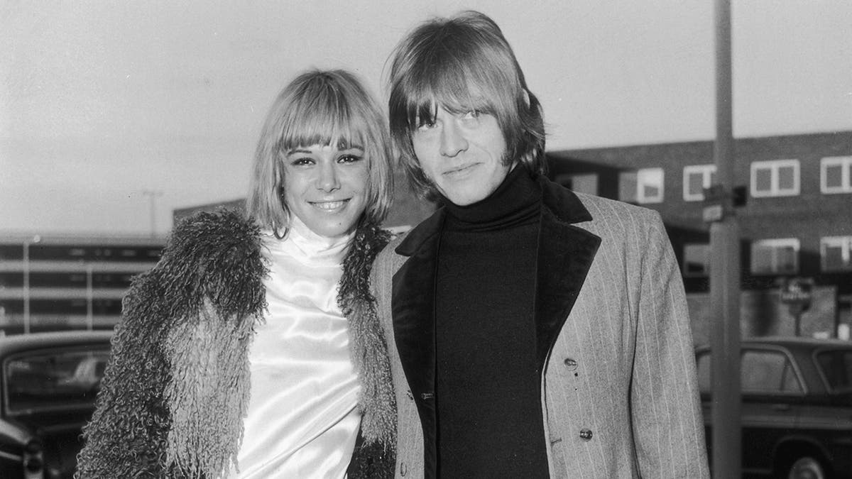 Anita Pallenberg smiling wearing a furry coat and white turtle neck dress posing next to Brian Jones who is also warmly dressed