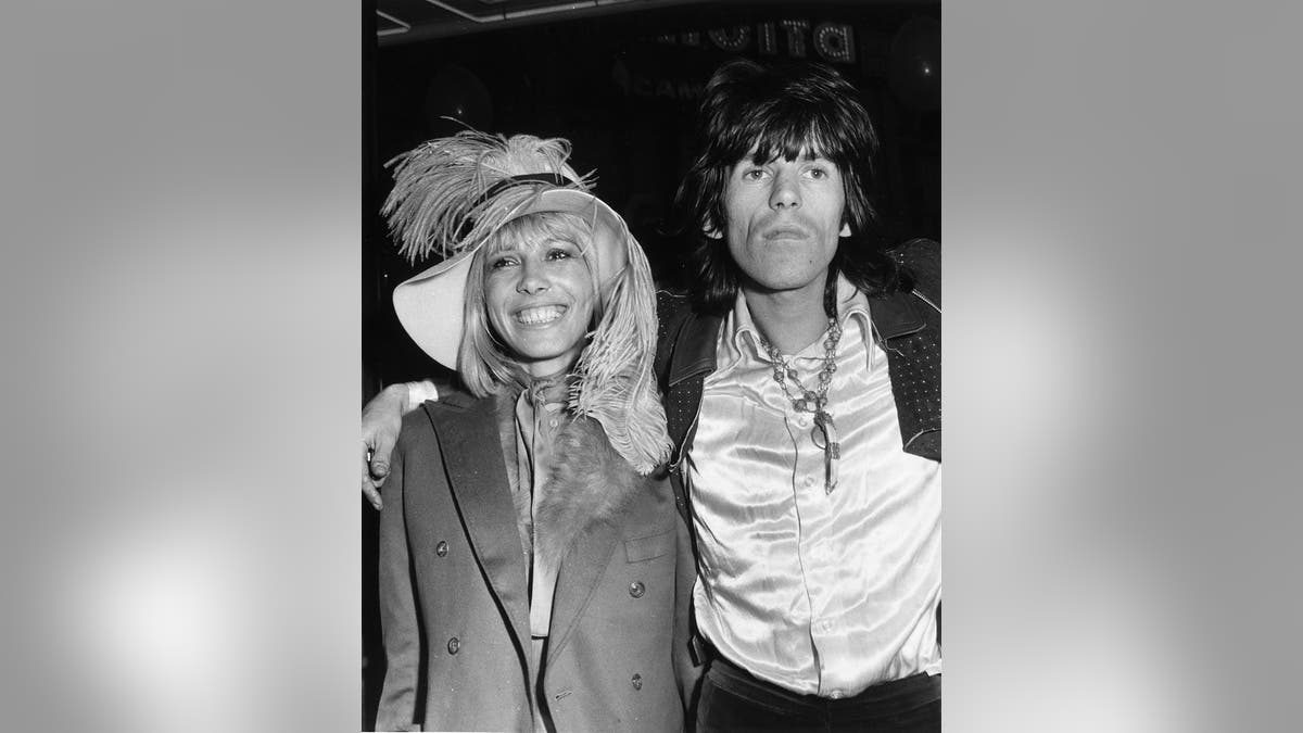 Anita Pallenberg wearing a peacoat and a feathered hat in the arms of Keith Richards