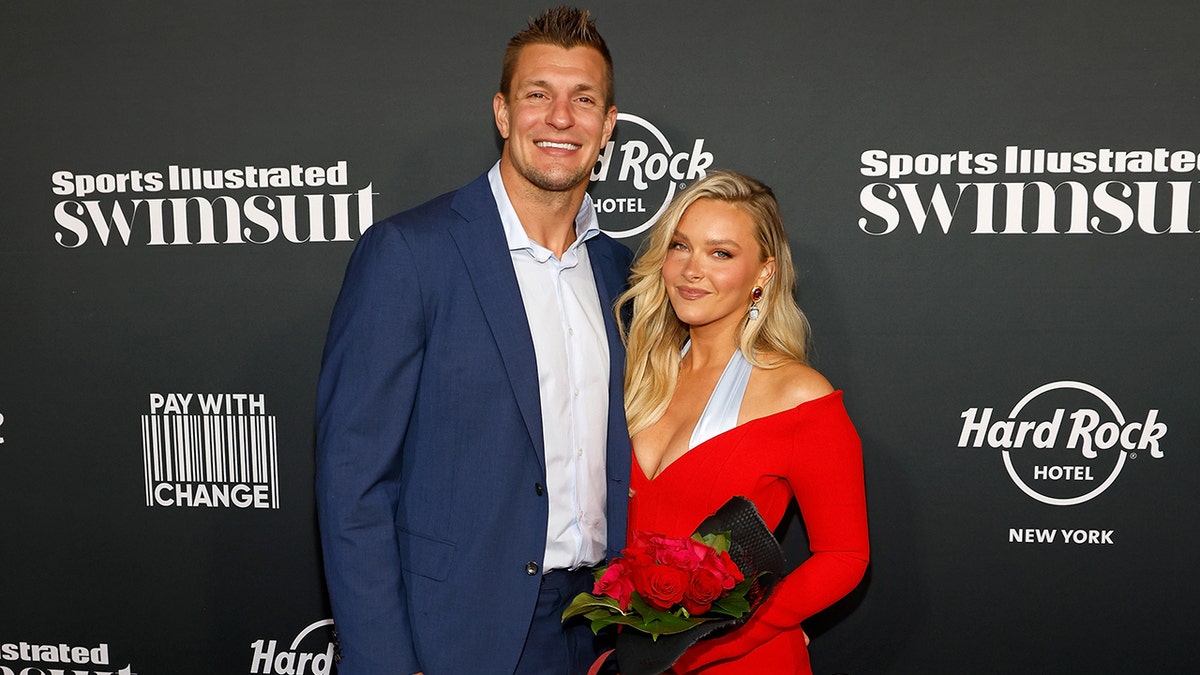 Rob Gronkowski wearing a blue blazer and a white shirt posing next to Camille Kostek wearing a red and blue dress