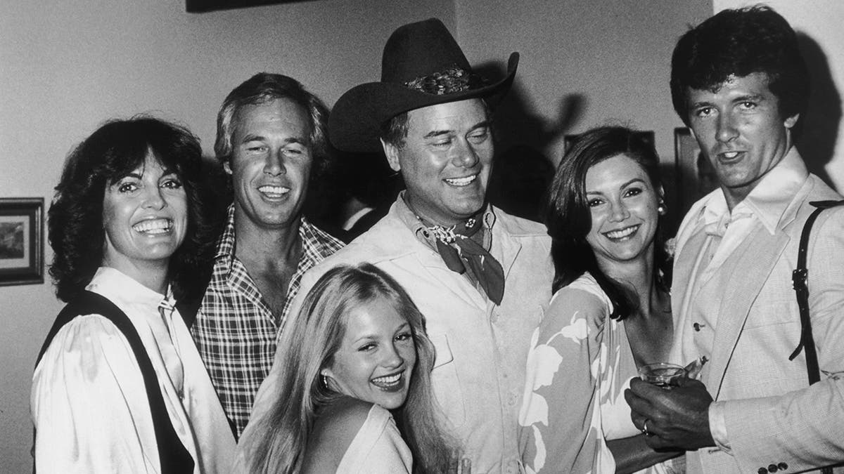 The cast members of Dallas in a black and white shot smiling with each other