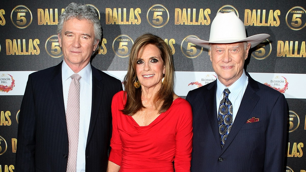 Patrick Duffy standing next to Linda Gray in a red dress and Larry Hagman in a dark blazer and cowboy hat