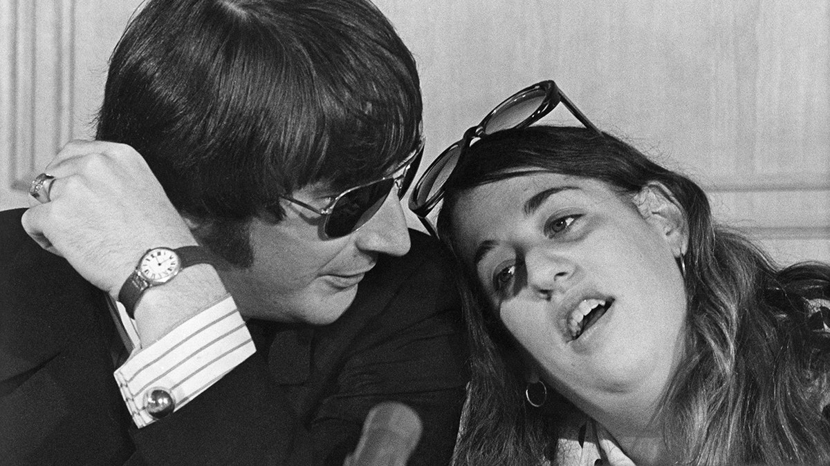 Cass Elliot leaning on Denny Doherty who is wearing a dark blazer, white shirt and sunglasses