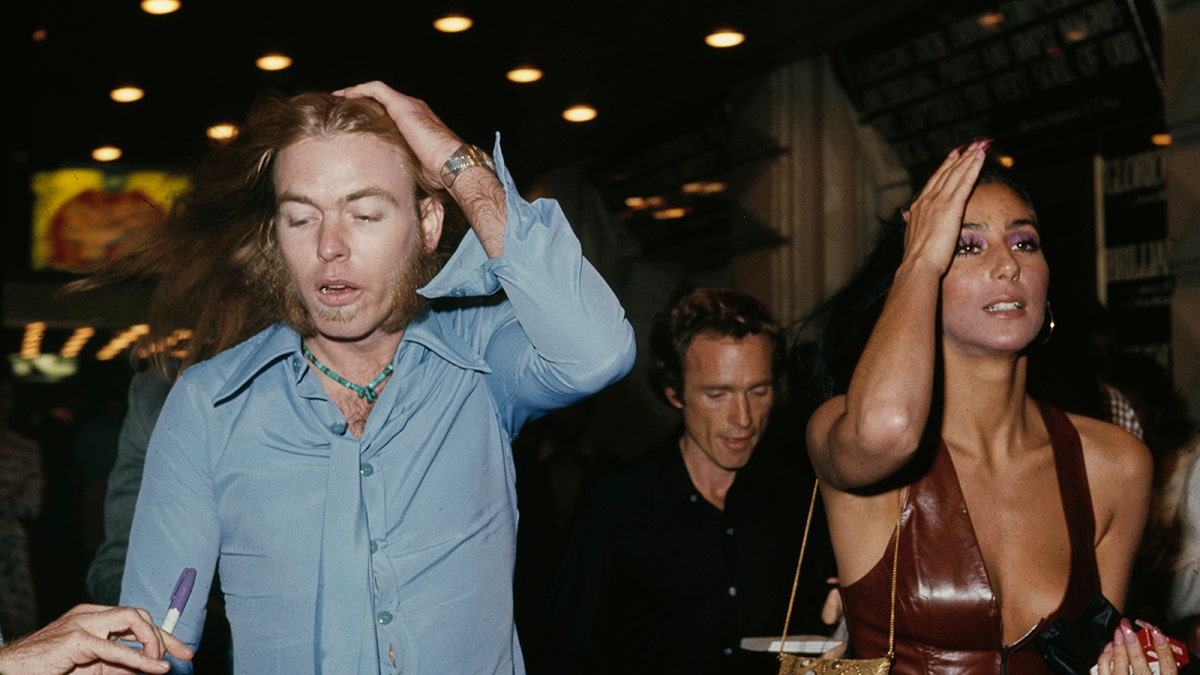 Gregg Allman wearing a light blue shirt walking next to Cher in a brown tank top