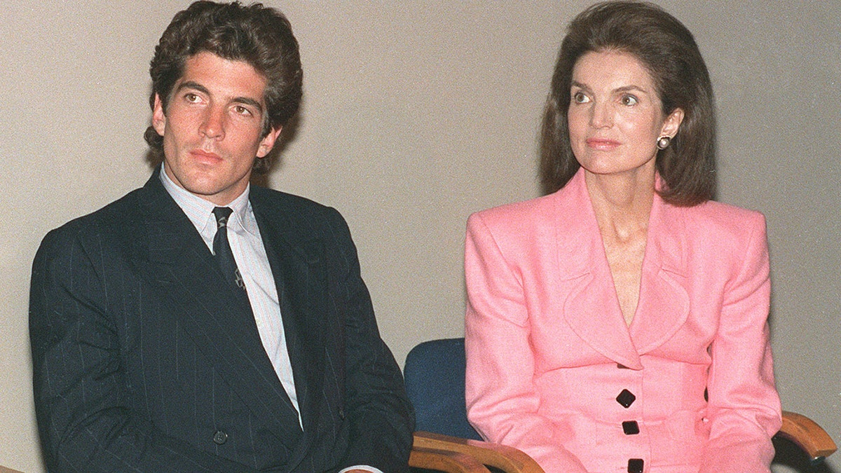 Jackie Kennedy wearing a bright pink suit sitting next to her son John F. Kennedy Jr. in a suit and tie