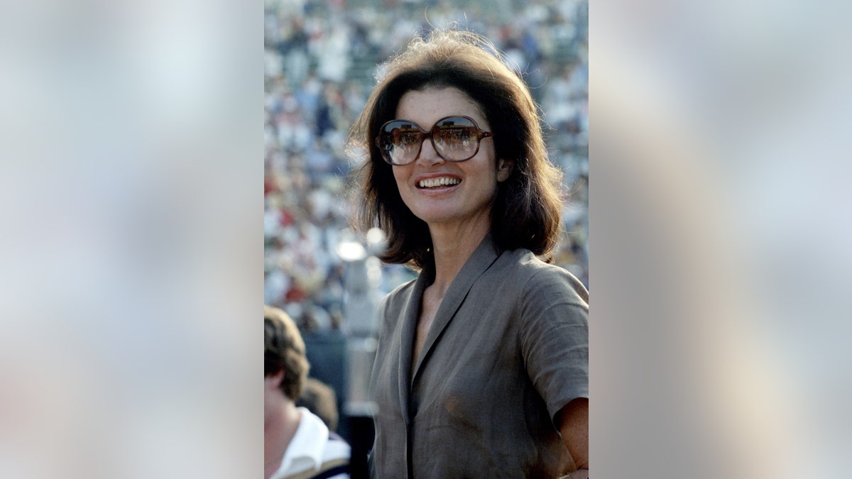 A close-up of Jackie Kennedy wearing her round sunglasses and a grey suit