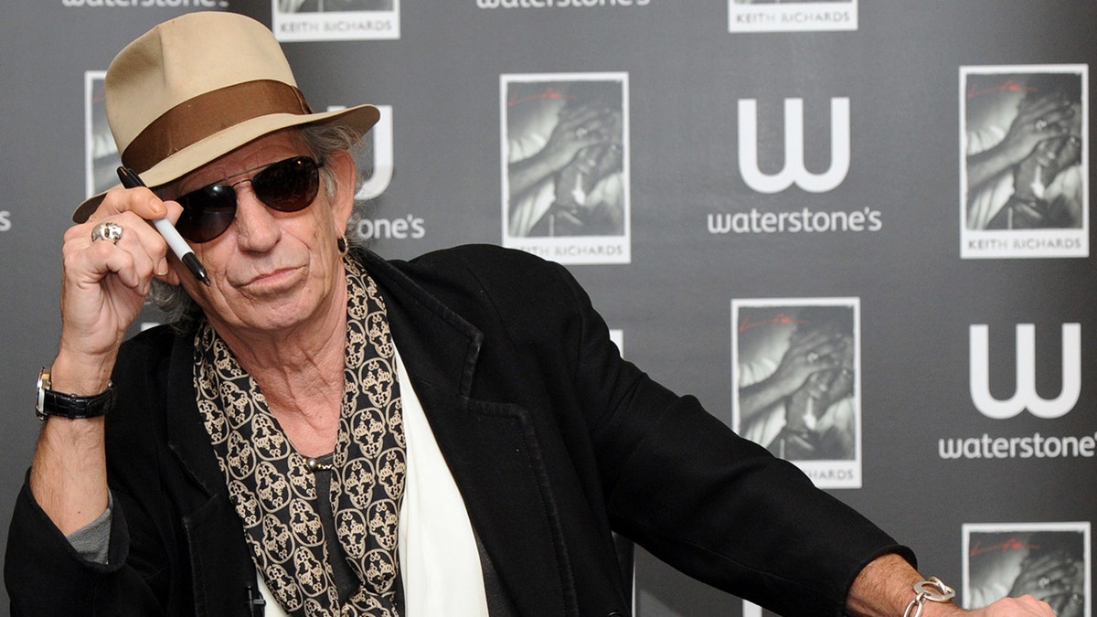 Keith Richards wearing a black jacket, white shirt, scarf and a fedora hat with sunglasses while holding a pen