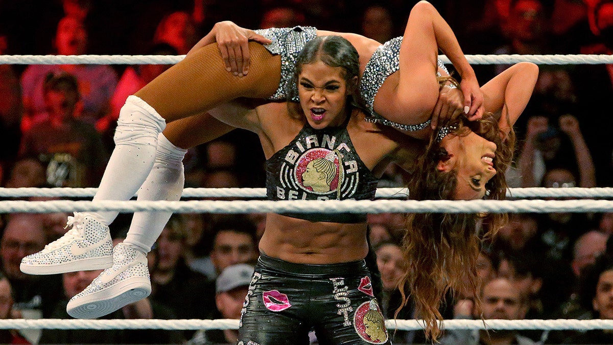 Leah Van Dale in a black and white two piece being lifted by Bianca Belair in the ring