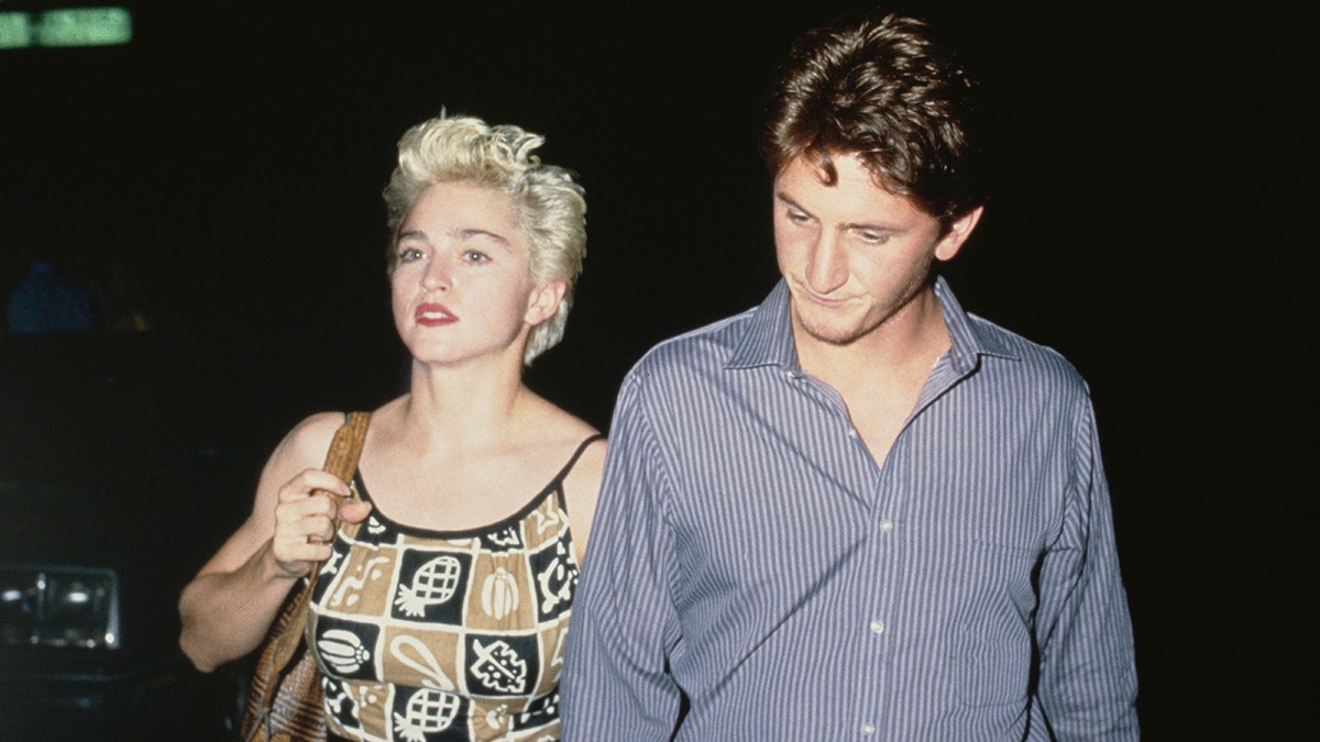 Madonna usando um vestido multiestampado andando ao lado de Sean Penn em uma camisa azul cinza