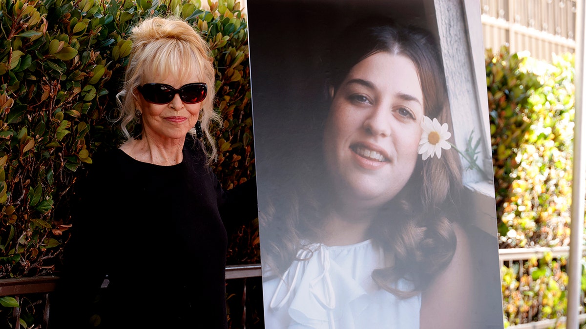 Michelle Phillips wearing a black dress and sunglasses standing next to a portrait of Cass Elliot