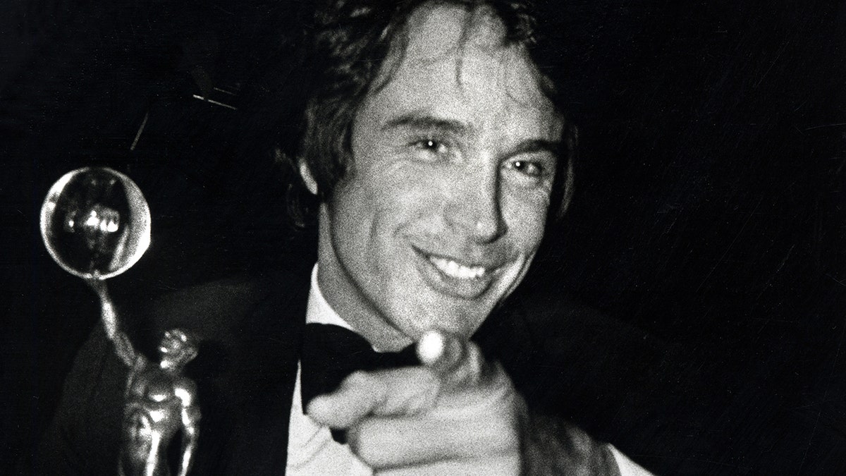 Warren Beatty holding an award and pointing at the camera wearing a tux