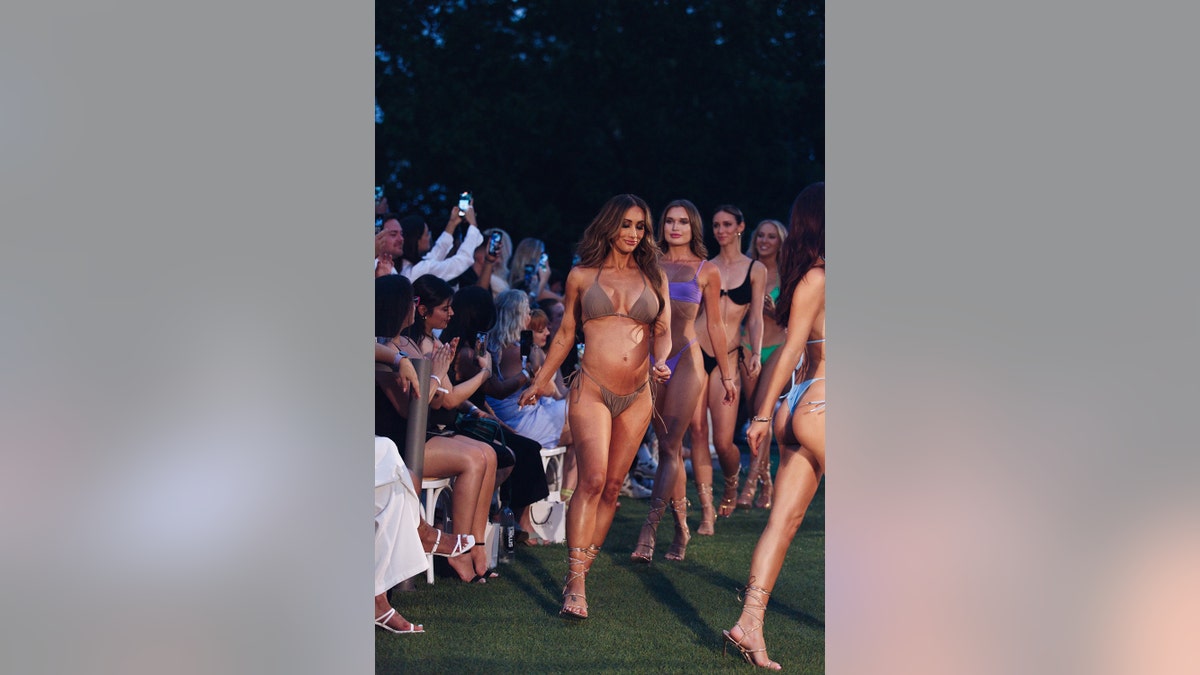 Leah Van Dale walking down the runway in a dark beige bikini with several women in swimsuits walking behind her
