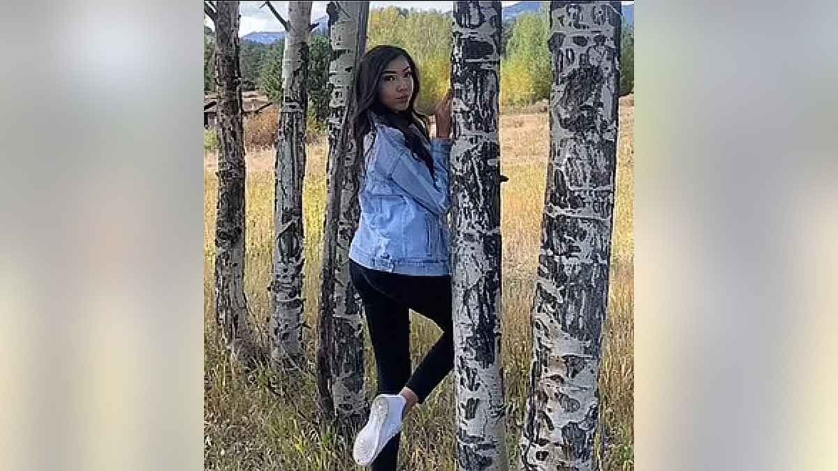 Colorado teen stands in between trees wearing a jean jacket.