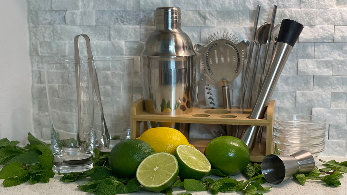 Barware kit with glasses, tongs, cocktail shaker, strainer, muddler spoons, and straws sit next to mojito ingredients.
