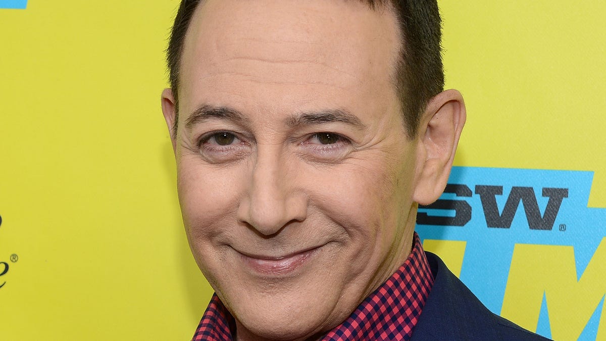Paul Reubens in a red checkered shirt and black jacket smirks at camera