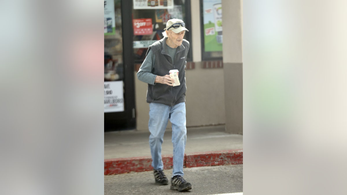 Gene Hackman crosses the street wearing baseball hat and sunglasses.