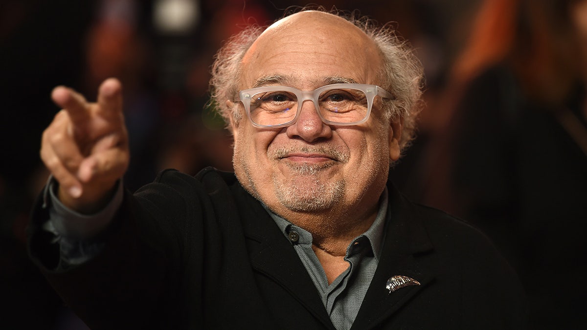 Danny Devito smiles and puts out a peace sign towards the crowd on the carpet