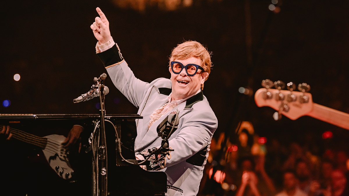 Elton John sitting at piano and pointing up