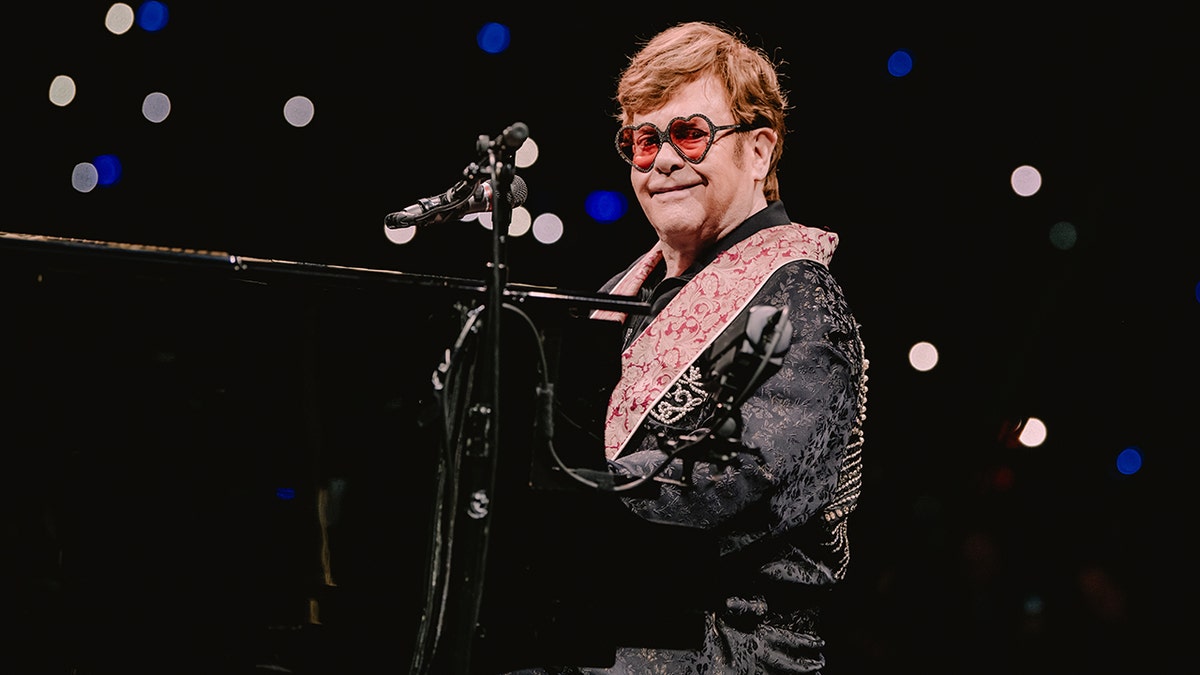 Elton John sits at piano smiling wearing sunglasses