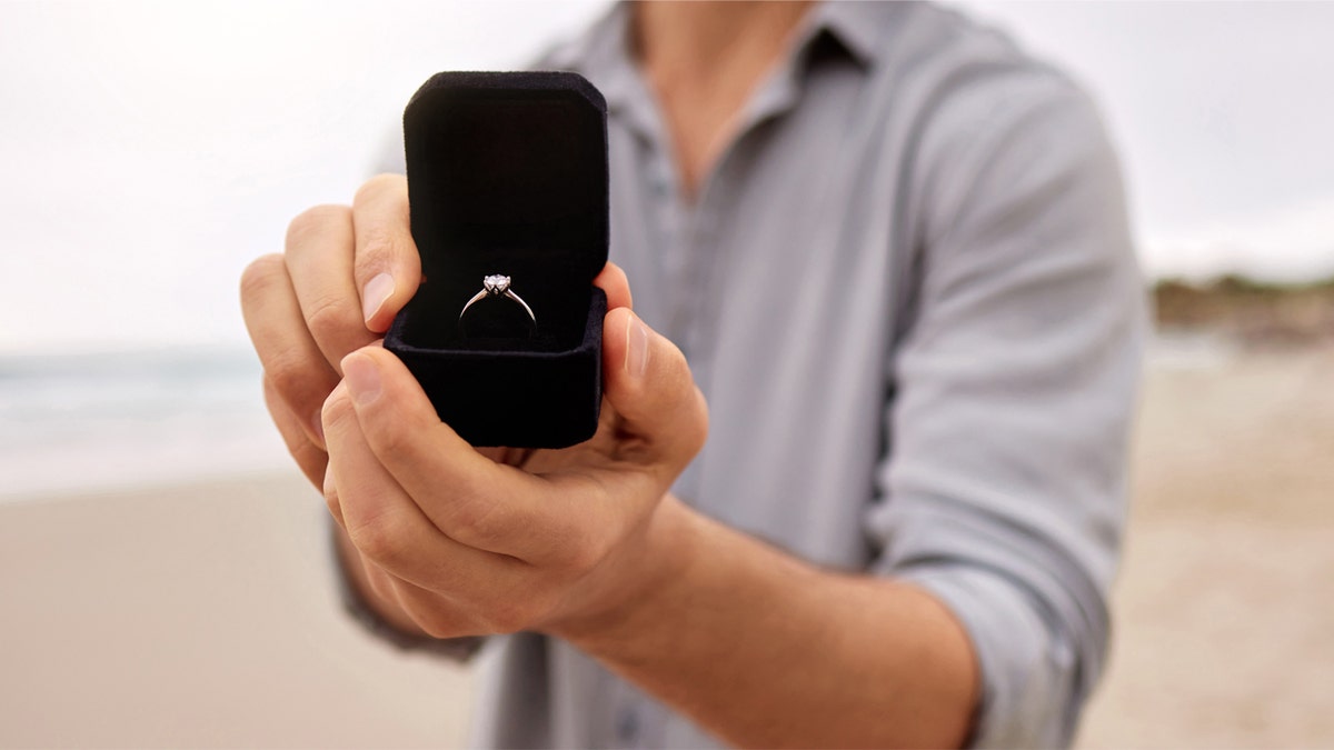 Shot of a man on a beach holding an open jewelry box with an engagement ring.