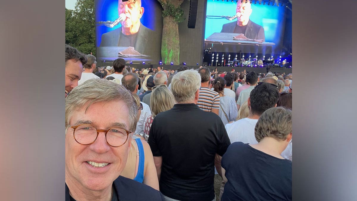 Greg Palkot smiles in selfie with Billy Joel performing behind him 