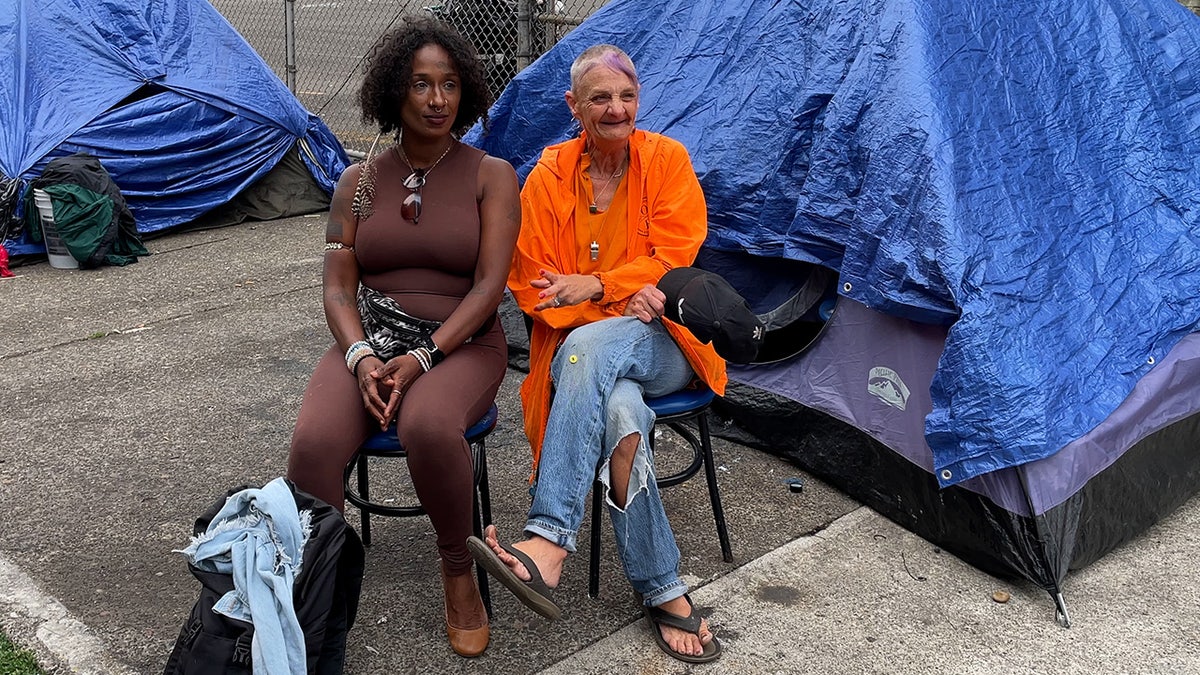 Two women sit on sidewalk in front of tents