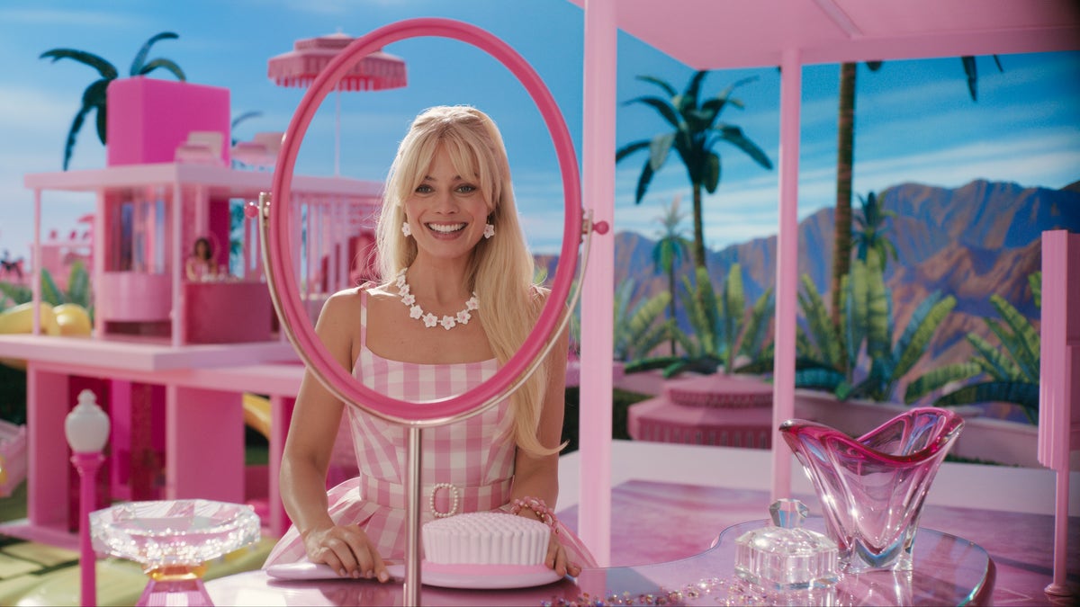 Margot Robbie em um vestido xadrez como Barbie no filme "Barbie" olhando através de um espelho sem vidro