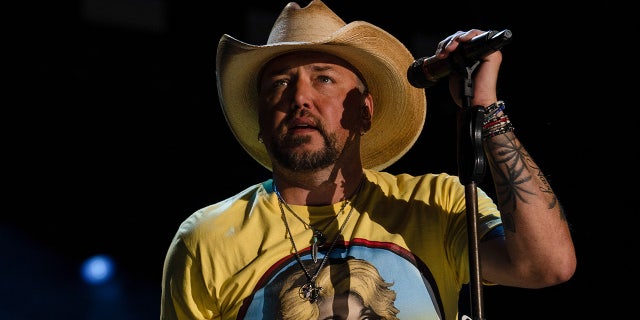 Jason Aldean in a yellow shirt with Dolly Parton on it and a tan cowboy hat holds the microphone stand