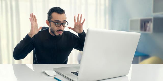 Man looking shocked, throwing hands in air while looking at laptop