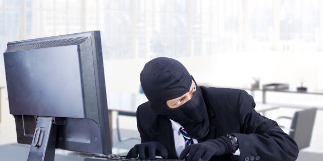 Hacker in black suit, gloves, and mask, typing on computer, looking to the right