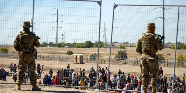 Migrants wait in line adjacent to the border fence