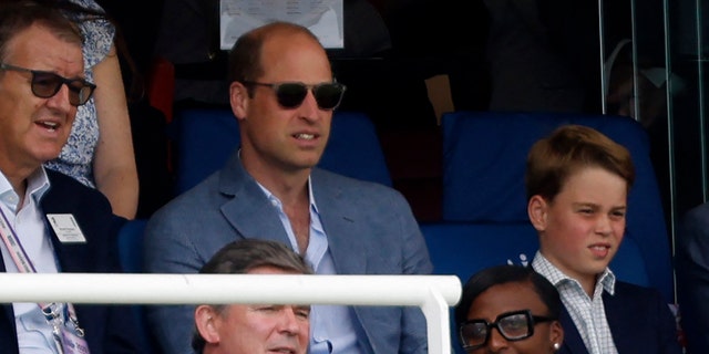 Prince William wearing sunglasses sitting in the stands next to Prince George