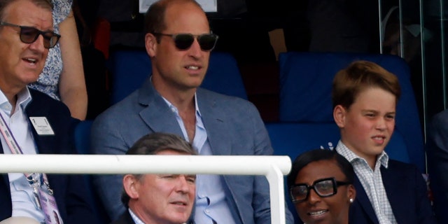 Prince William wearing sunglasses sitting in the stands next to Prince George