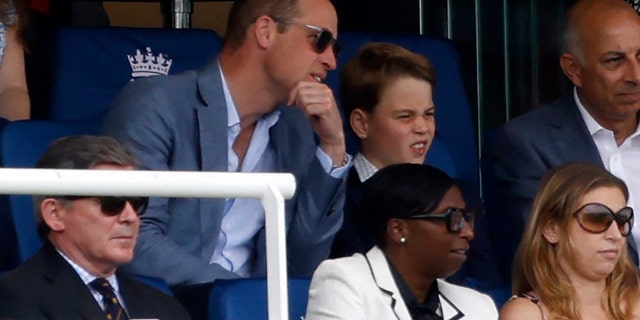 Prince William wears sunglasses and leans in to talk to Prince George in the stands