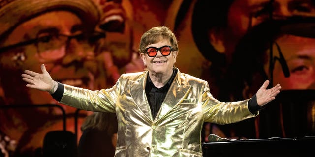 Elton John stands on stage in gold suit jacket with arms out.