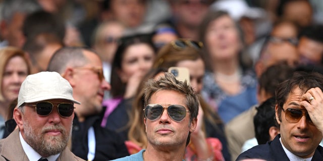 Brad Pitt wearing sunglasses and blue shirt in the stands at Wimbledon.