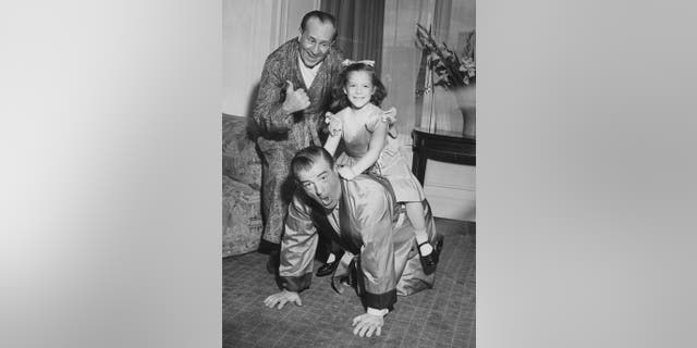 Abbott and Costello in suits and smiling at the camera as they play horse ride with daughter Chris Costello