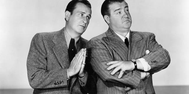 Bud Abbott wearing a suit with praying hands as Lou Costello in a suit and tie looks away