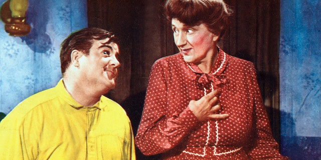 Bud Abbott wearing a polka dot burgundy dress and wig as a woman with Lou Costello wearing a yellow shirt while admiring him