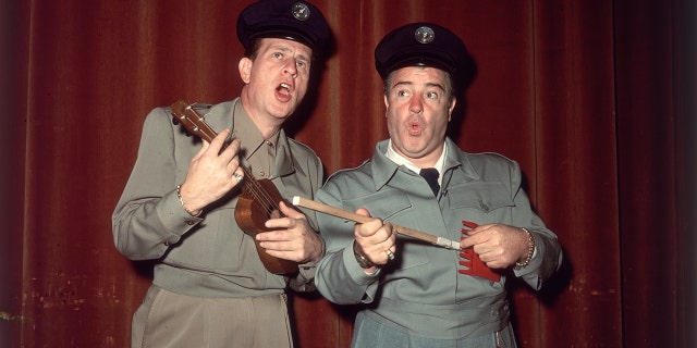 Bud Abbott holding a ukelele and singing with Lou Costello holding a gardening tool as a guitar