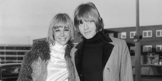 Anita Pallenberg smiling wearing a furry coat and white turtle neck dress posing next to Brian Jones who is also warmly dressed