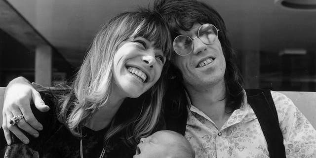 Anita Pallenberg smiling holding her son next to Keith Richards wearing sunglasses