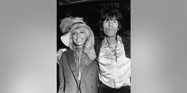 Anita Pallenberg wearing a peacoat and a feathered hat in the arms of Keith Richards