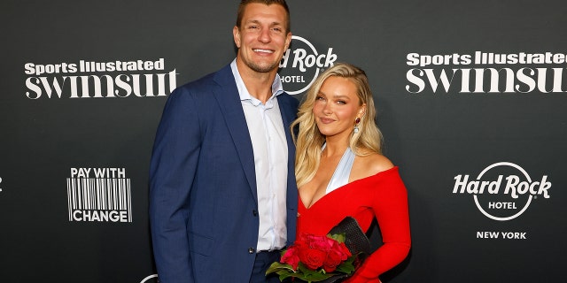 Rob Gronkowski wearing a blue blazer and a white shirt posing next to Camille Kostek wearing a red and blue dress